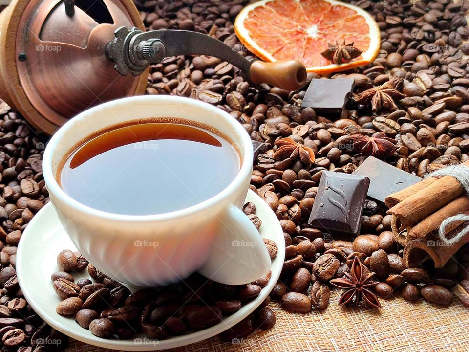 Coffee. There are coffee beans on the burlap. In the foreground is a white cup of coffee. On the coffee beans are cinnamon sticks, slices of dark chocolate, star anise, dried orange and a lid from a manual coffee grinder