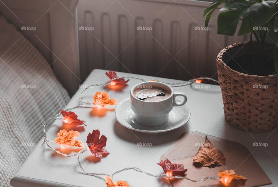 One cup of hot coffee with a garland of burning autumn leaves on the bedside table by the bed in the bedroom early in the morning, close-up side view.