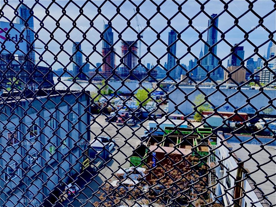 “Long Island City” and in the furthest distance “Manhattan” can be seen through this metal mesh fence that protects pedestrians from falling when they cross the “Pulaski Bridge”. 2023. Hypnotic Productions