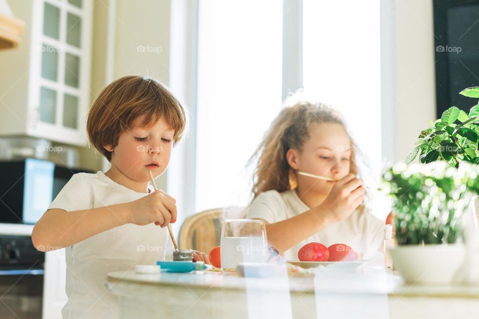 Two siblings brother and sister toddler boy tween girl painting easter eggs on kitchen at home on the spring sunny day