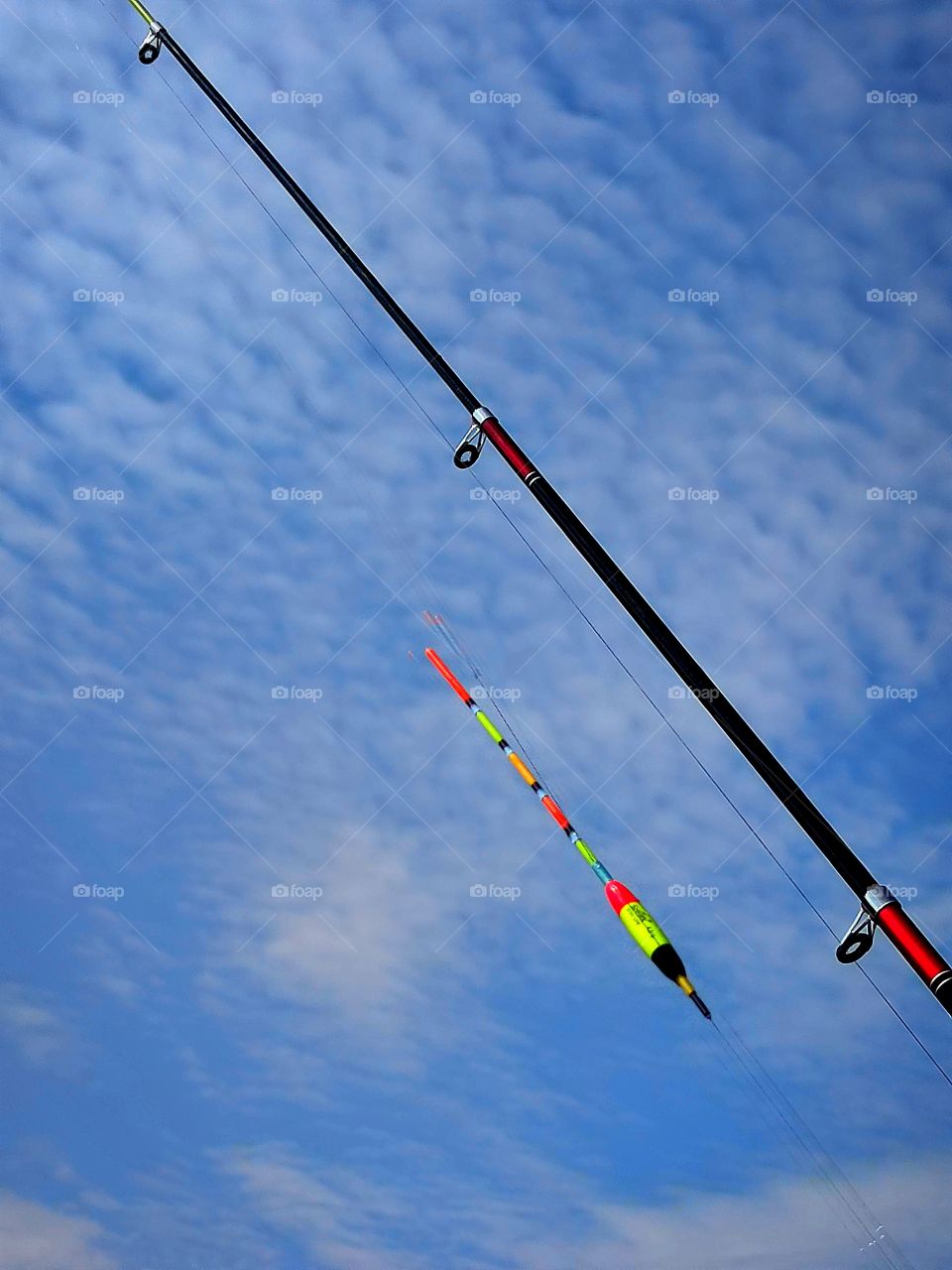fishing rod, fishing line and multicolored float against a blue sky with white clouds