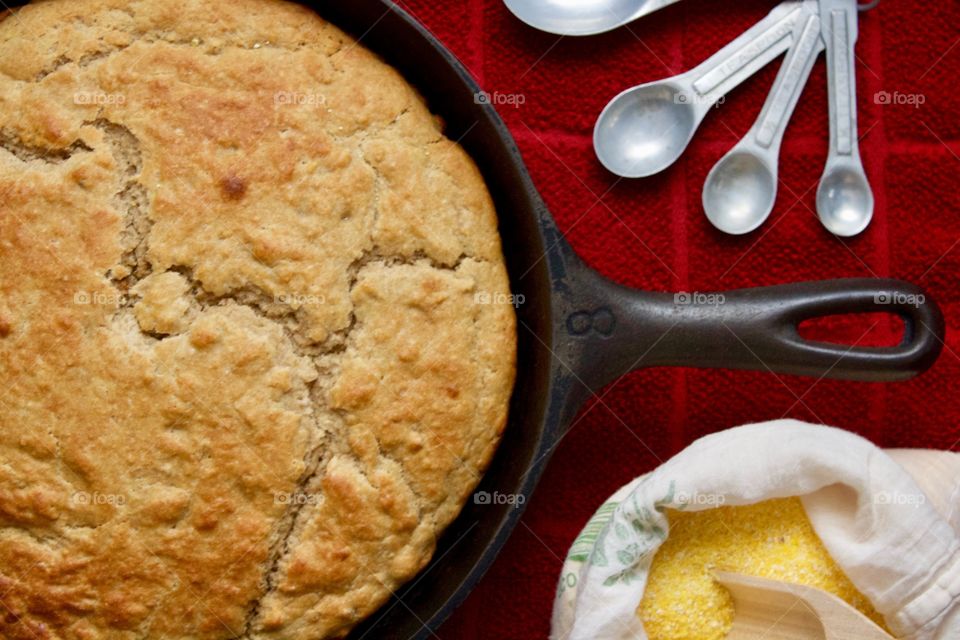 Closeup flat lay of sourdough cornbread in a cast iron skillet, cornmeal and wooden scoop in a flour sack, vintage measuring spoons on a red kitchen towel