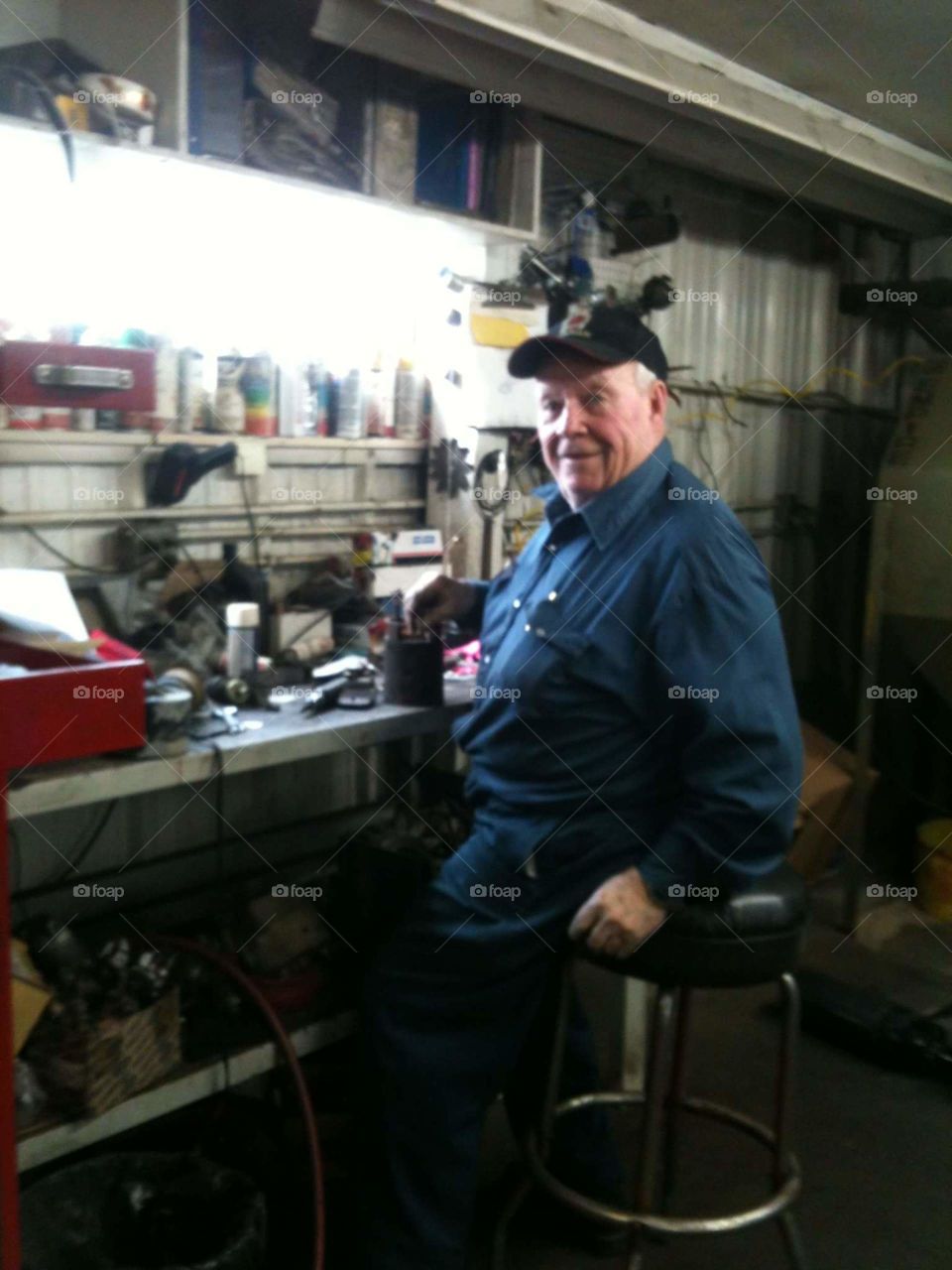smiling old man sitting on stool at his workbench taking a break for a pose. he is comfortable in his element.