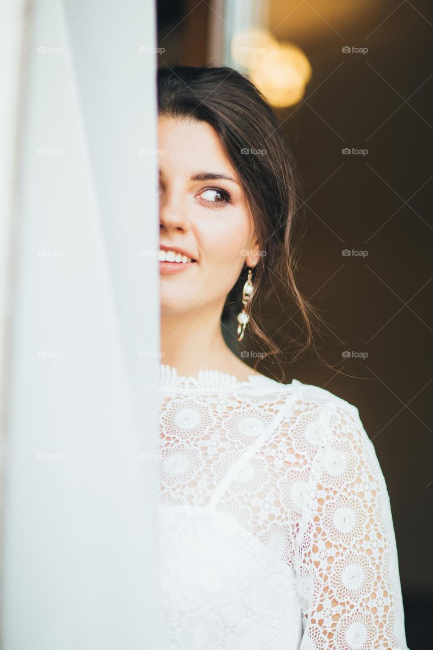 Beautiful smiling bride brunette young woman in the white lace dress near window, close up