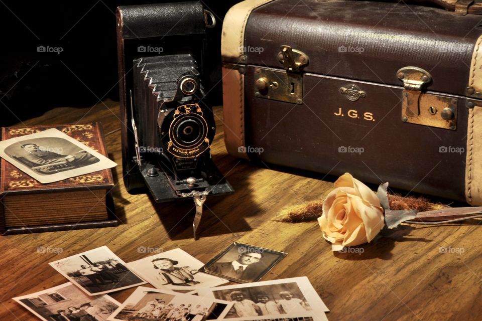 Old fashioned camera beside an old piece of luggage with very old photos scattered on the table still life