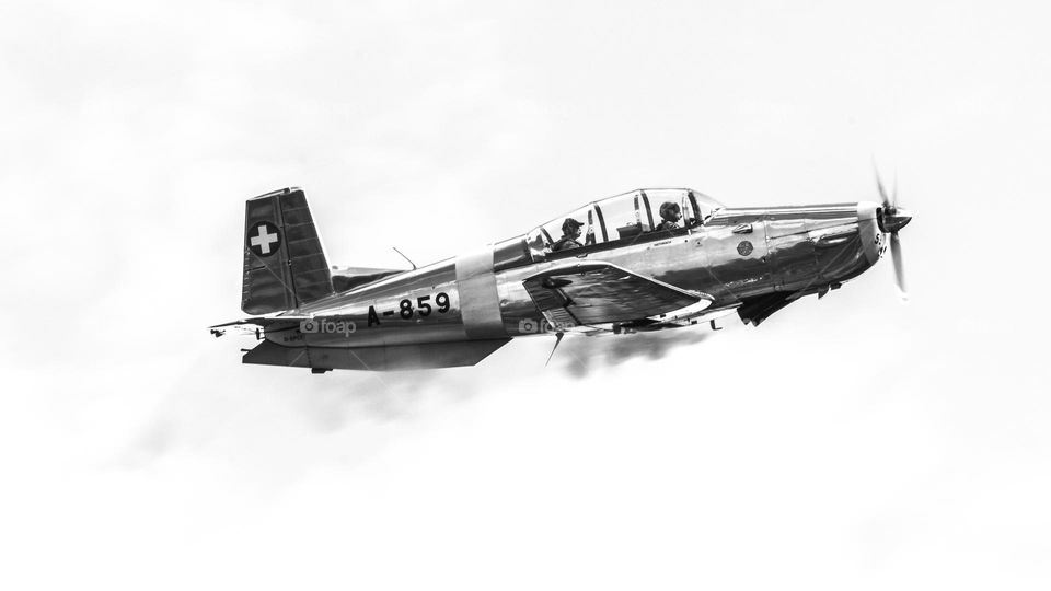 A black and white side Portrait of a vintage stunt airplane letting smoke loose to make a trail. the two airmen are sitting behind eachother and are very focussed.