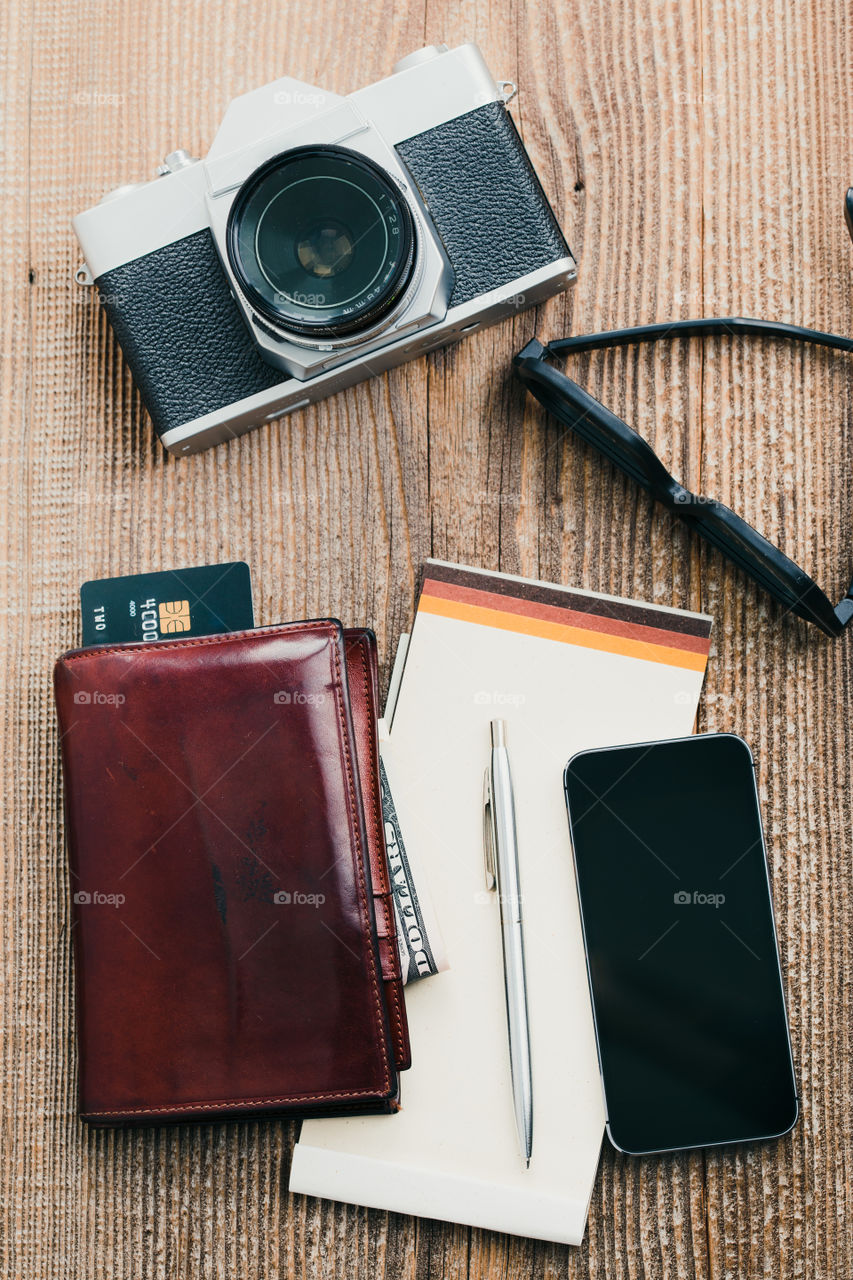 Smartphone with blank screen, camera, wallet, dollar banknotes, debit credit cards and notebook on wooden table. View from above. Portrait orientation