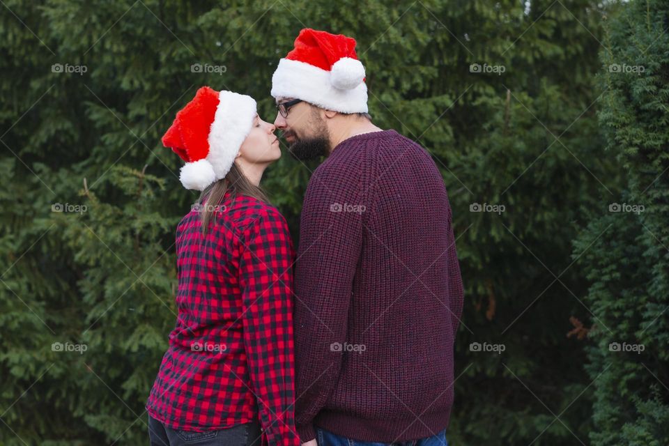 Young beautiful loving couple stands on the street near the Christmas trees meets Christmas hugs and kisses in a Santa hat and red sweaters