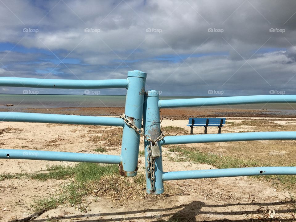 Marine blue gate closed and locked with chain and padlock barring vehicles from accessing beach with blue bench and ocean horizon in the background