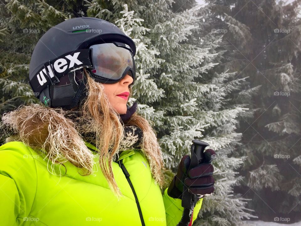 Portrait of woman wearing Uvex ski glasses and Uvex ski helmet and holding ski poles with pine cone trees covered in snow in the background 