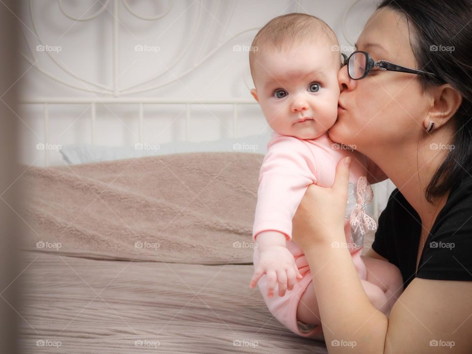 Beautiful side view of a Caucasian woman mother gently kissing her little baby daughter on the cheek while holding her with her hands on the bed in the bedroom during the day.