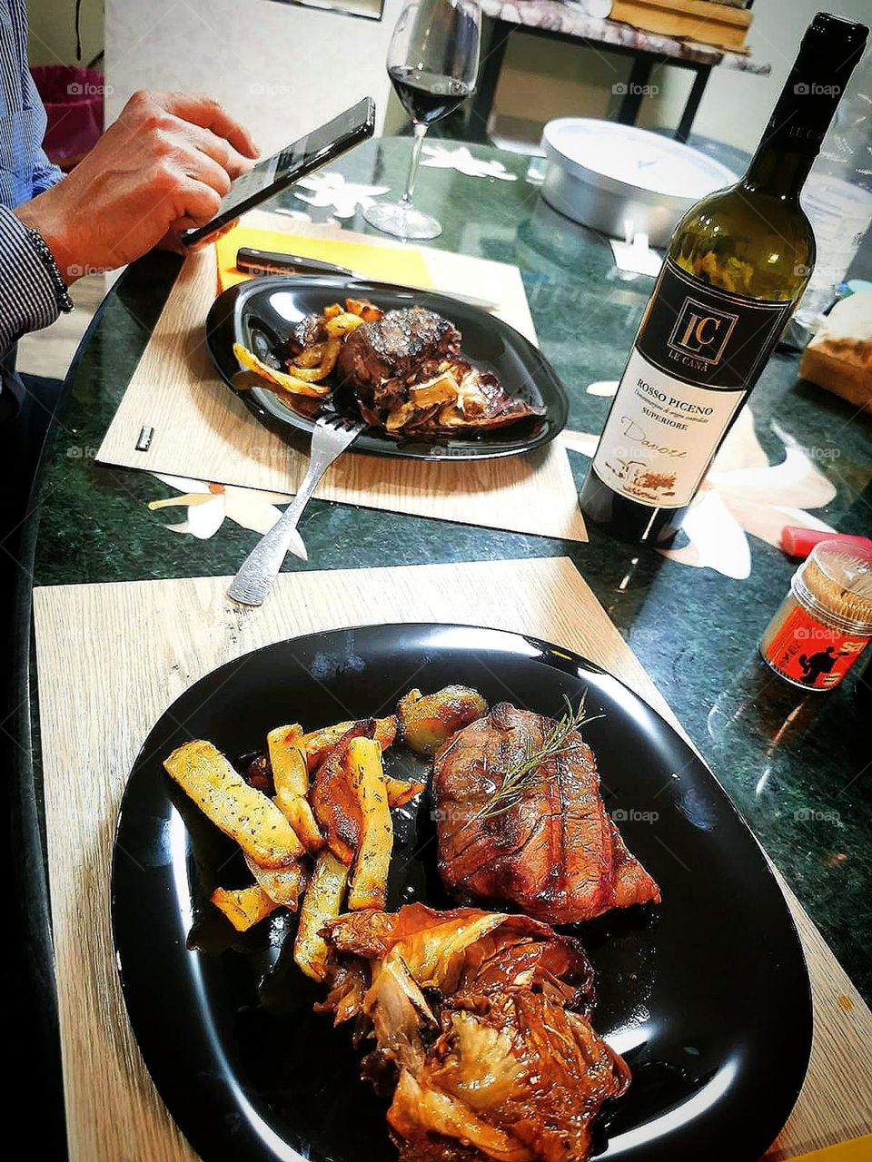 Food. On the table are two rectangular black plates. Each plate contains fried and stewed meat and fried potatoes. Stand on the table are: a bottle of wine, a glass of wine. At the second plate, man's hands with a telephone are visible