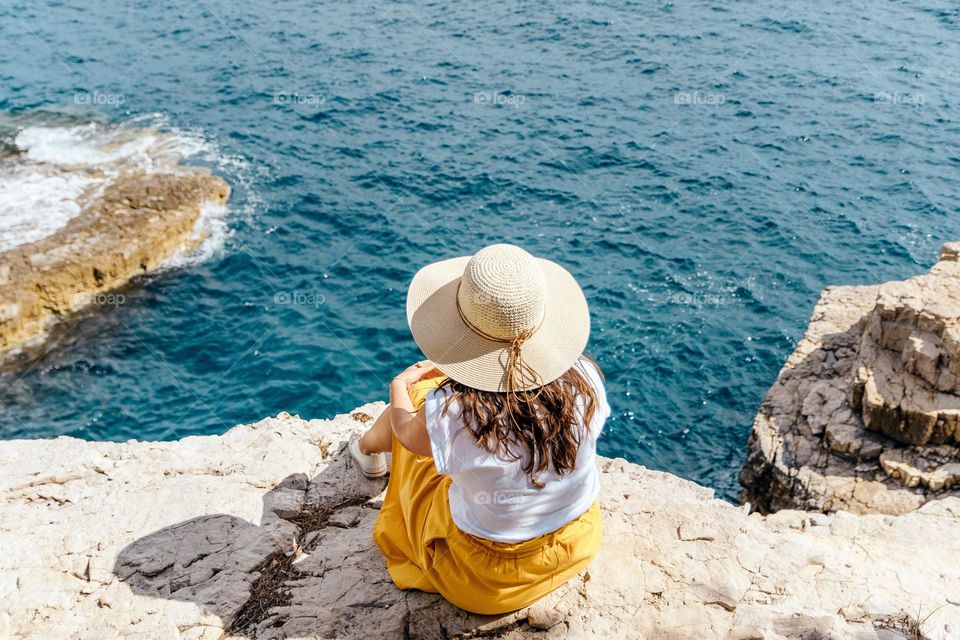 Rear view of young woman wearing summer clothes and sun hat relaxing on cliff above sea