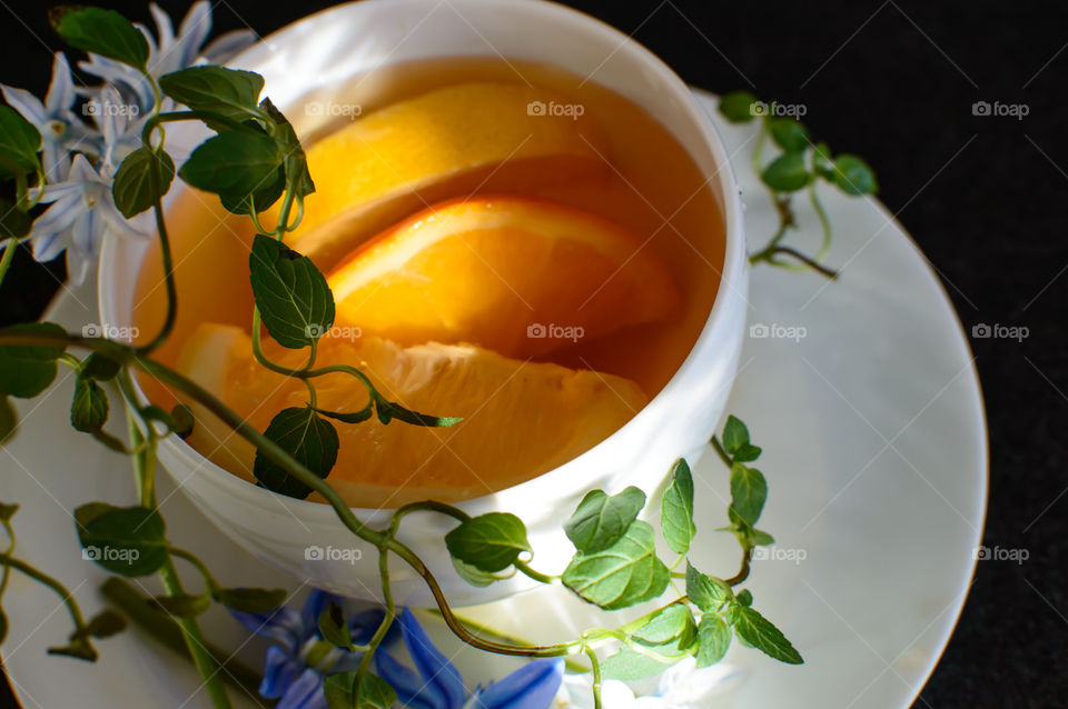 A beautiful morning cup of tea with fresh sliced orange and lemon fruit surrounded by fresh growing mint leaves on vines and lily flowers 