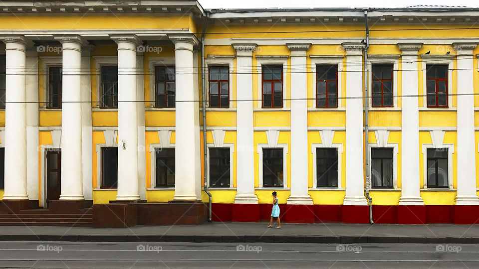 Young woman wearing a summer blue dress using mobile phone and walking by the old yellow building in the city 