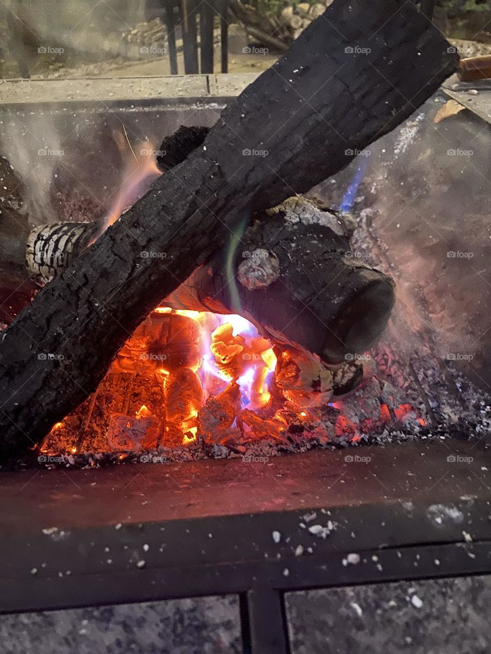 This is a photo of a campfire with burning logs. The embers and flames show a mix of red, orange, and blue colors, indicating the high temperature and presence of different chemicals.