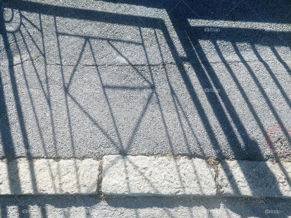 The shadow of the fence on the concrete sidewalk.  Representation of geometric shapes: circle,  triangle, and rectangle