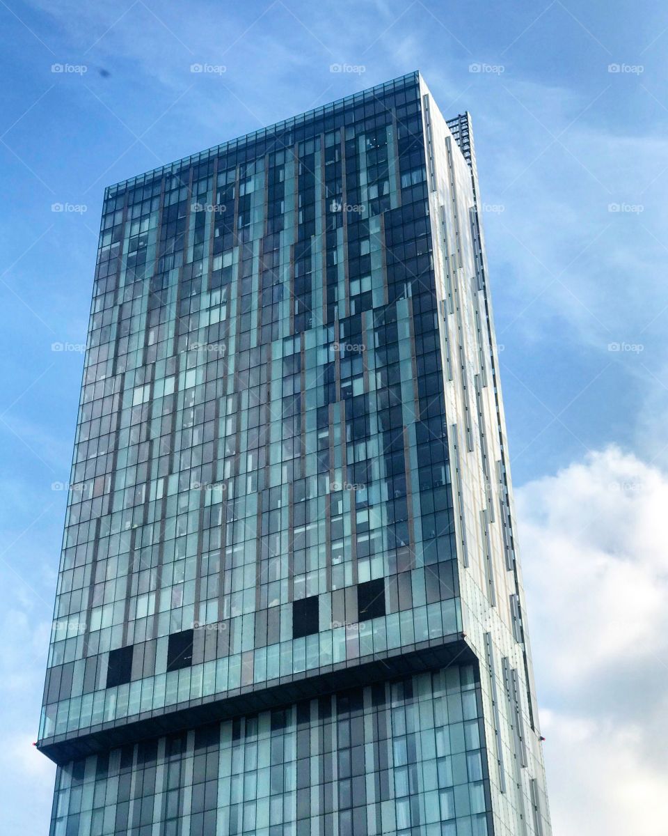 Hilton Hotel in Manchester, captured in a glorious sunny and clear day. The building stands tall, and is bathed in a blue glow from the sky and it’s glass filled architecture