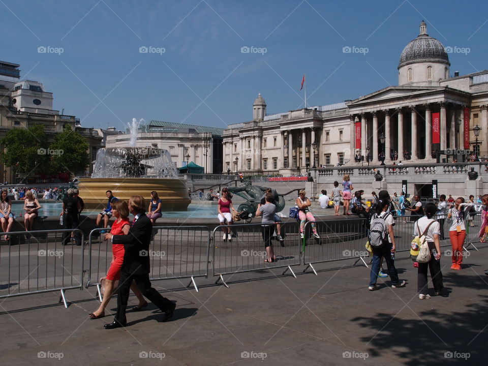 Crowds of people walk in an open square with a beautiful fountain in London on a sunny summer day. 