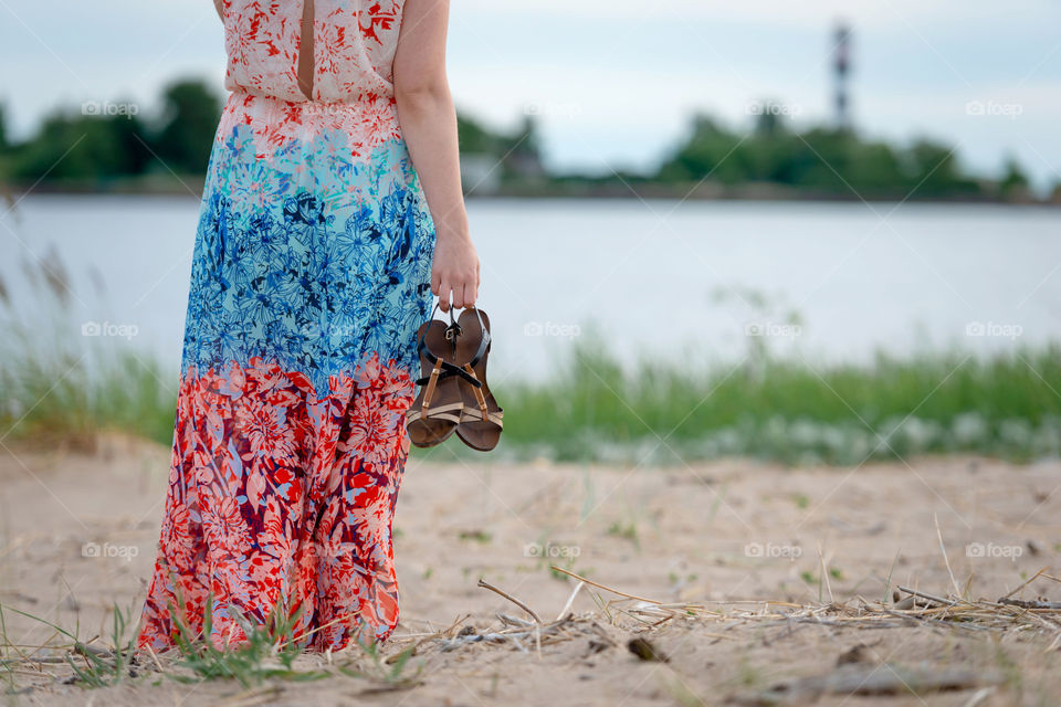 A young woman wearing a colorful long dress and shoes in her hands standing on the beach and looking at the lighthouse. View from the back.