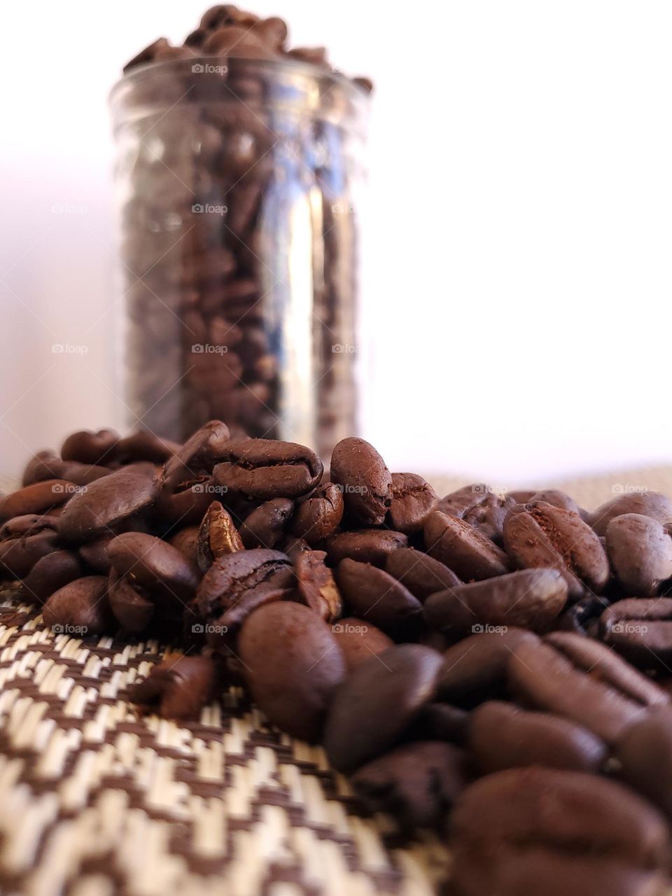 A clear glass jar of coffee beans in the background and spilled coffee beans directed towards the camera.