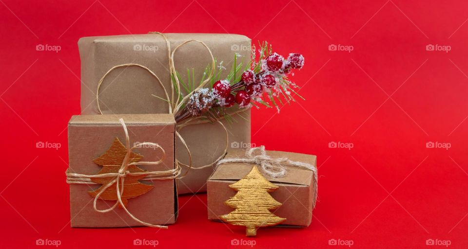 Three homemade gift boxes tied with jute thread with spruce branches and decorative Christmas trees lie on the left on a red ion with copy space on the right, close-up side view. Holiday gifts concept.