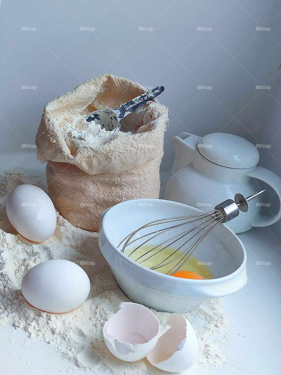 White flour is scattered on a white surface.On the flour are two white eggs,two white eggshells.Nearby is a white bowl with a broken egg and a metal whisk.In the background is an open cloth bag of flour, which contains a black spoon.