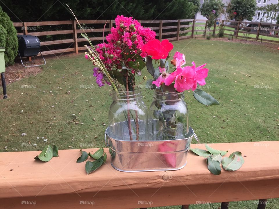 Two glass jars full of pink flowers placed in a metal tub.  Roses and lilacs.  Leaves strewn around.  Outside grass.