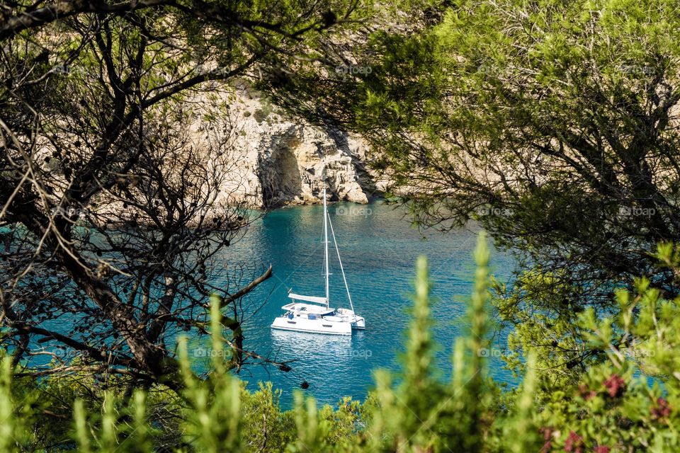 A yacht anchored in the blue sea of Cala llonga bay, Ibiza. Framed by trees.