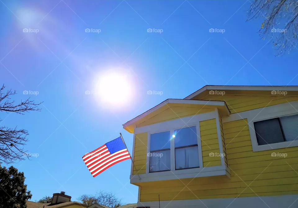 USA flag on a yellow residential home on a clear blues blue sky day and the sun out.