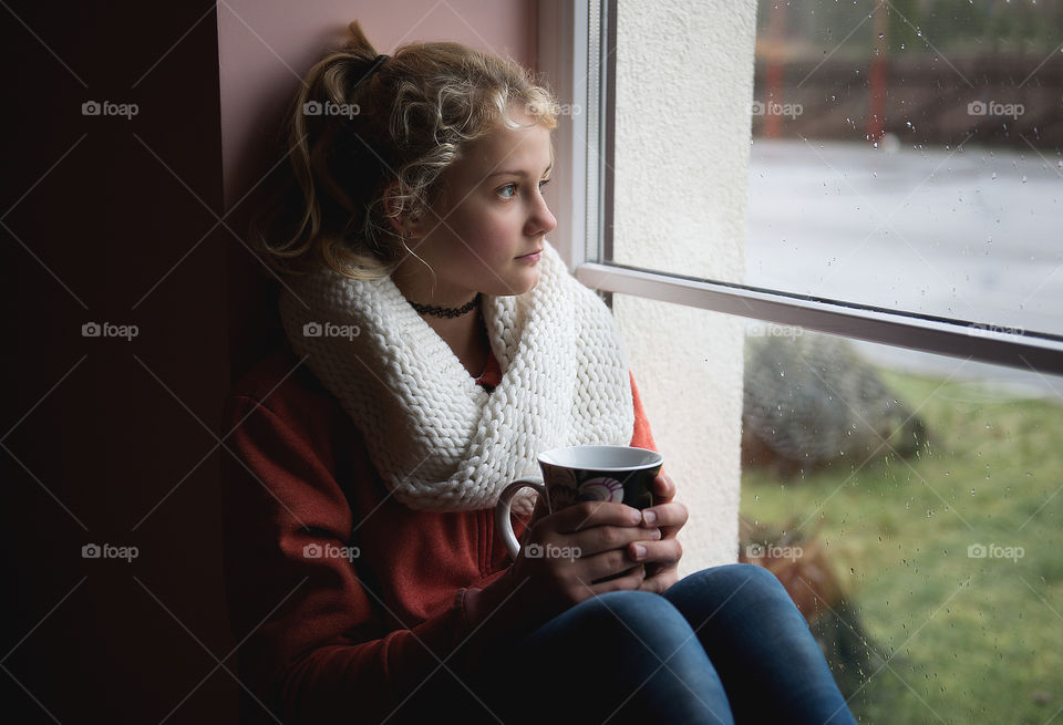 A girl with a cup of tea looks through the window as it rains in the autumn