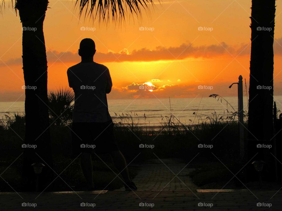 Sun peeking through the clouds during an exceptional sunrise in New Smyrna Beach, FL. Vivid orange and yellow sky and large sun as man watches from back yard. Great shadows of him and palm trees as sun rises in background. Calm Atlantic Ocean on a clean and clear day, July 1, 2017 by Rank180, LLC.