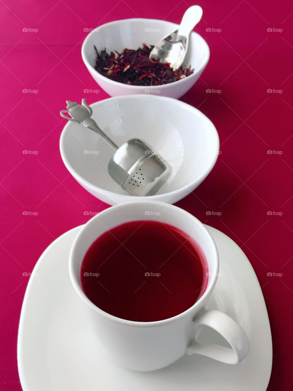 Tea Time - vertical arrangement of hibiscus tea in white cup, stainless steel teapot steeper in white bowl, dried hibiscus flowers in white bowl on magenta fabric background 