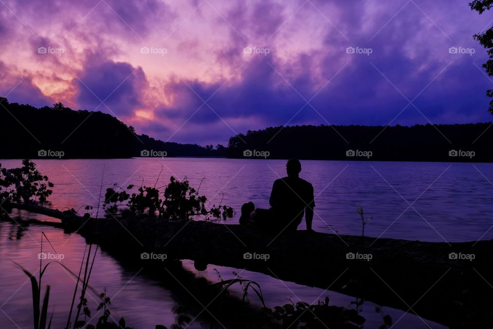 Silhouette of a man leisurely resting on a log over the water watching the cloudy sunrise at Lake Johnson Park in Raleigh North Carolina. 
