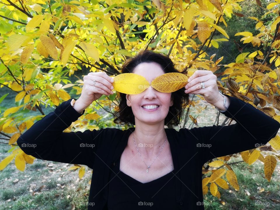 smiling woman puts yellow autumn leaves in front of her eyes