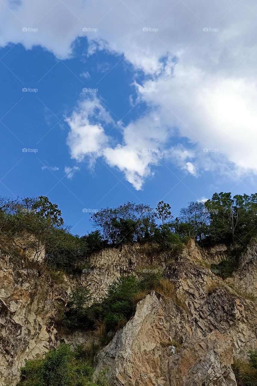 rocky cliff with vegetation at the top and blue sky with white clouds