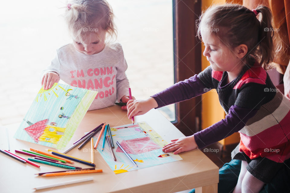 Two little girls drawing a colorful pictures of house and playing children using pencil crayons standing at table indoors