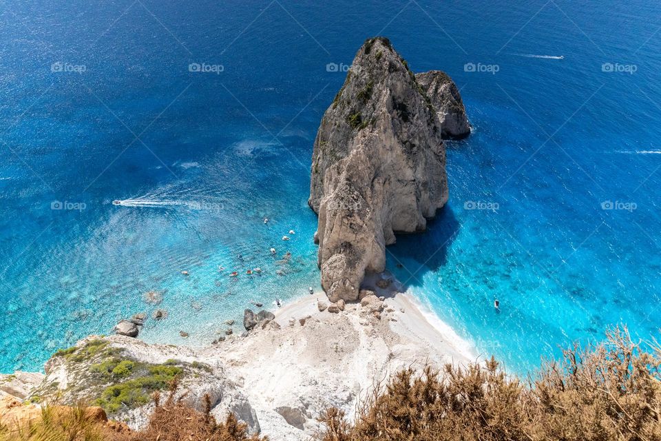 Beautiful aerial view of a solitary rock in the blue clear sea near the shoreline with boats passing nearby, close-up view from above.