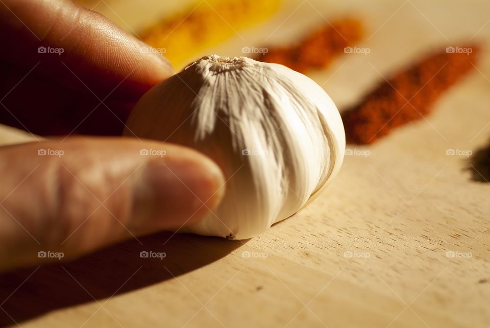 A closeup of a man holding a clove of garlic with plentiful and colorful herb spices in the background in preparation for a healthy meal.