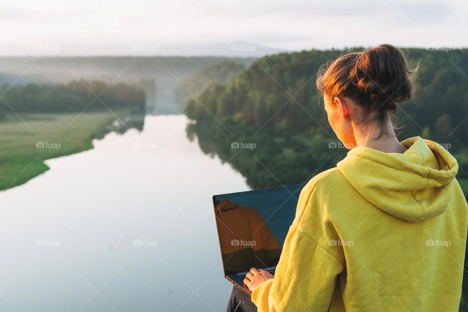 Young woman traveler freelancer in yellow hoodie with opened laptop on beautiful view background