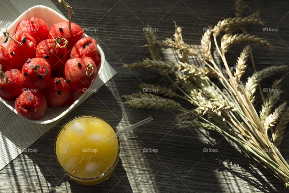 Cooling, orange juice with ice, red watermelon and ears of wheat are on a wooden table in the sun.