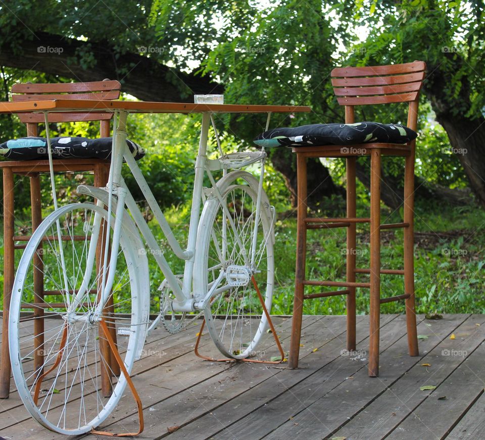 One lonely white bicycle with a wooden board on the top and two chairs.  Bicycle as a table