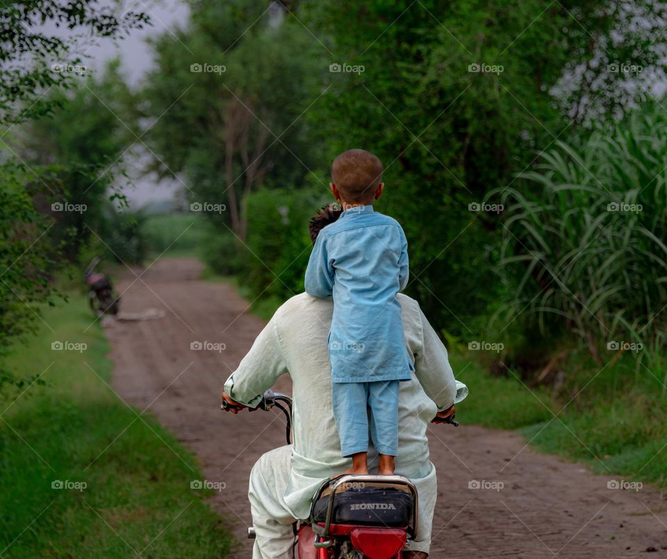 This image depicts a rural scene with a father and son riding a motorcycle along a narrow, brick-paved path surrounded by lush greenery. The man, dressed in light-colored traditional attire, drives the bike, while the young boy,