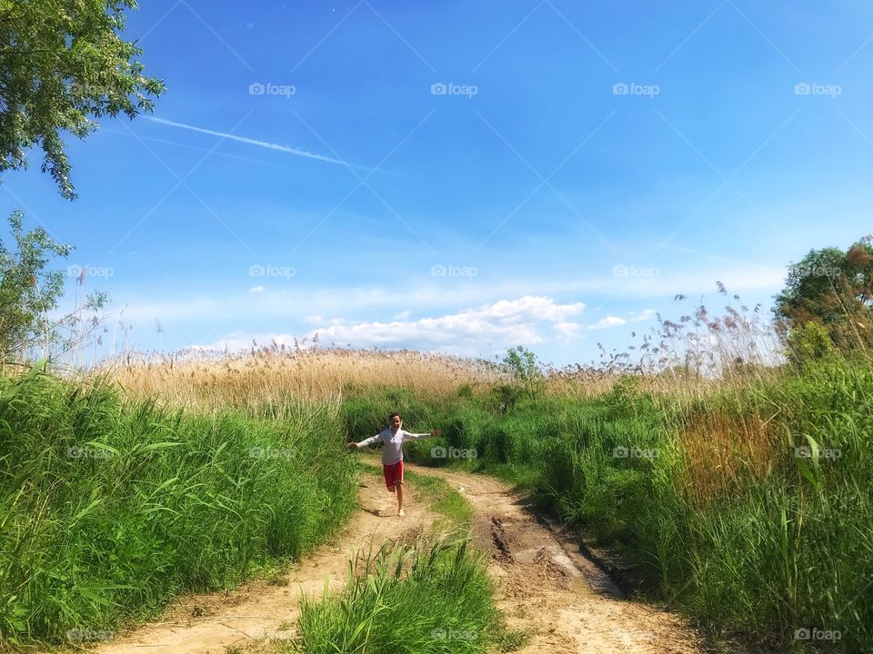 a boy in a white shirt runs spread his arms like wings along a deserted country road, joy, happiness, ease, field, green grass, village, holidays, summer, blue sky, sun, clear sunny day