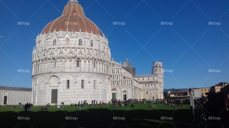 Photo of the monument of Piazza dei Miracoli in Italy, with a great blue sky with no cloud and lot of persons in the meadow under the monument. On background the leaning tower of pisa