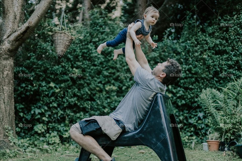 Handsome young Caucasian dad with brown curly hair in work uniform throws up his little daughter while sitting on a garden long chair in the backyard garden of his house after work, close-up side view. The concept of fathers, dad love, at home,