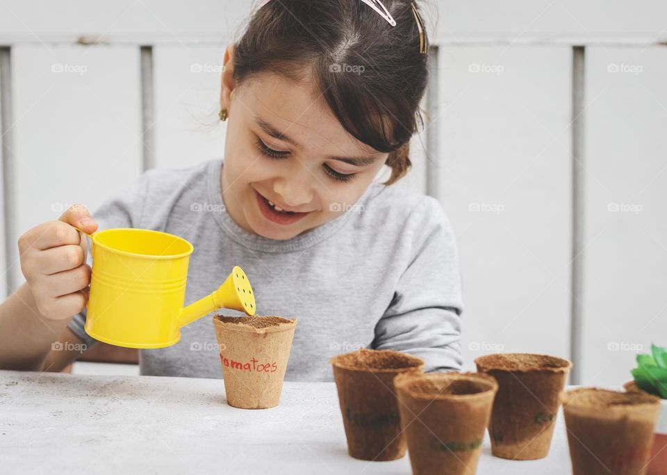 A beautiful little caucasian brunette girl with a cute smile on her face waters the planted seeds in craft cardboard glasses with inscriptions with a yellow watering can, sitting at a light stone table in the backyard, close-up side view. The concept