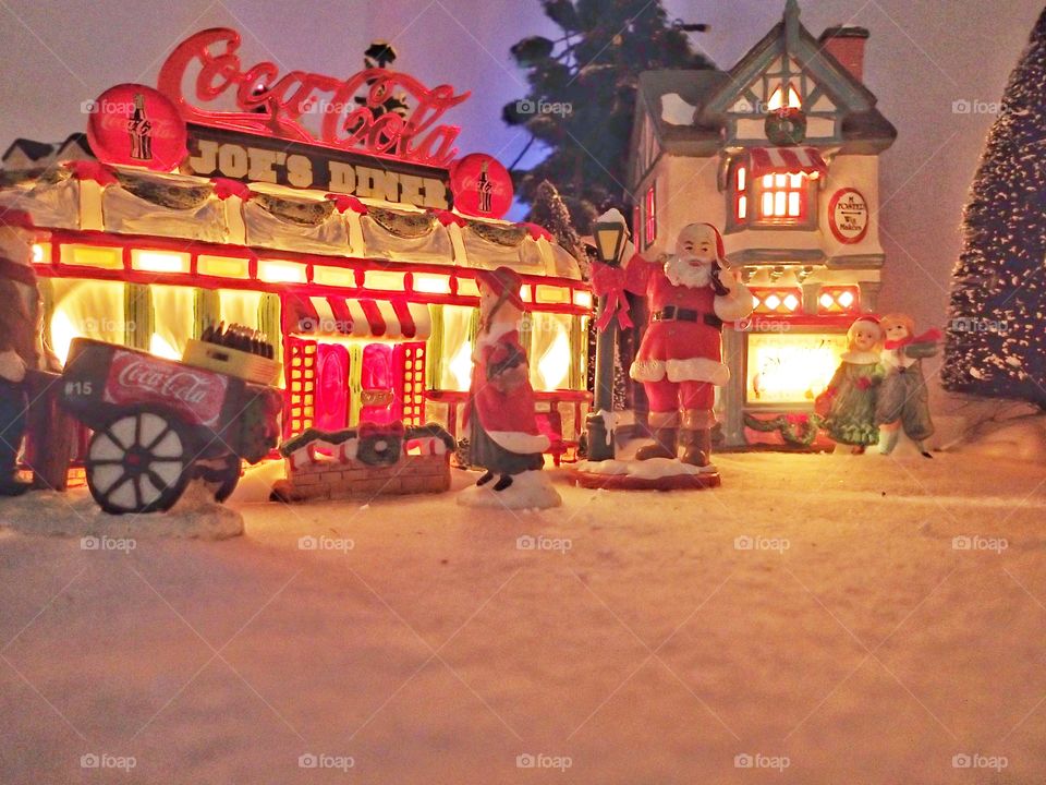 A Christmas village display with a Santa and other people in front of a diner with a big soda sign. 