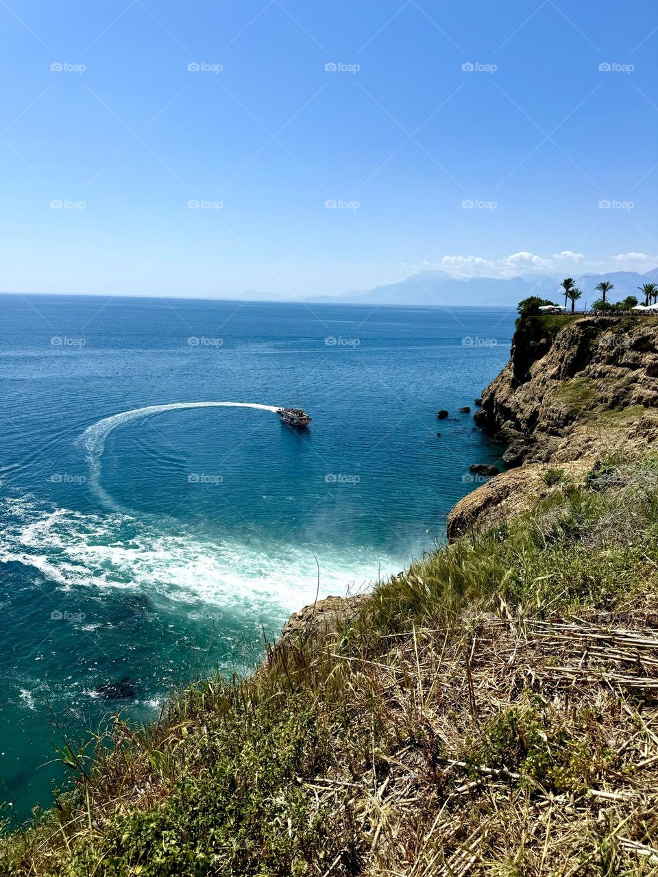 На фото изображен крутой обрыв, возвышающийся над  морем. Внизу, вдалеке, виден плывущий кораблик, оставляющий за собой белоснежный след на поверхности моря.