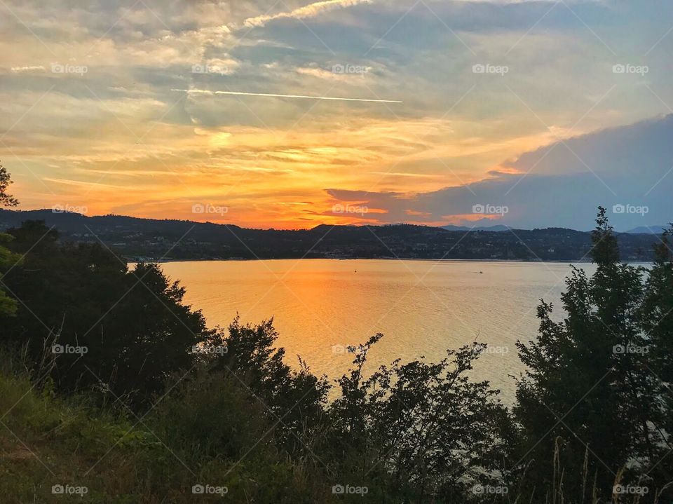 Sunset view . The surroundings have become dark, trees, bushes, mountains . Sky with clouds in orange, colors reflecting into the lake water surface . 
